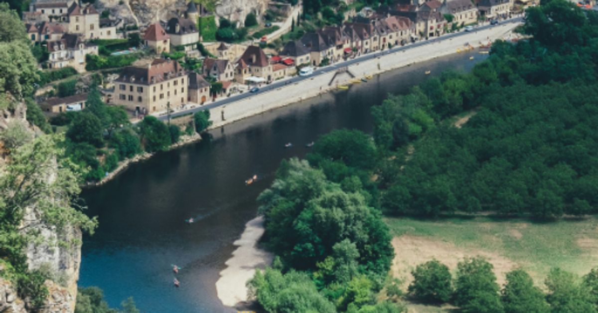 The Dordogne river winding through a medieval village built into limestone cliffs, surrounded by green forest
