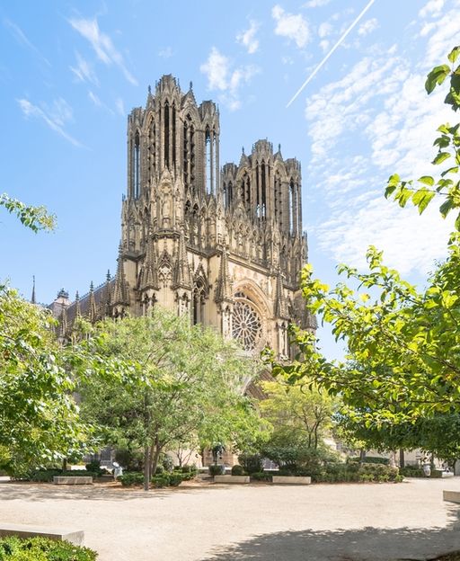 The Gothic facade of Reims Cathedral framed by green trees under a blue sky, Champagne region, France