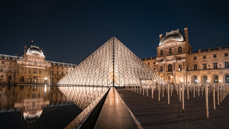Illuminated glass pyramid reflected in still water at night with classical buildings