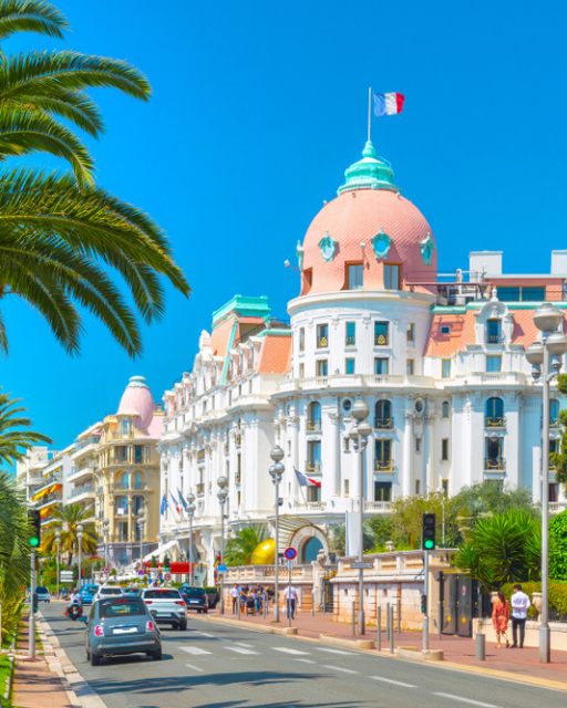 Belle Époque hotel with pink dome on tree-lined Mediterranean promenade under blue sky