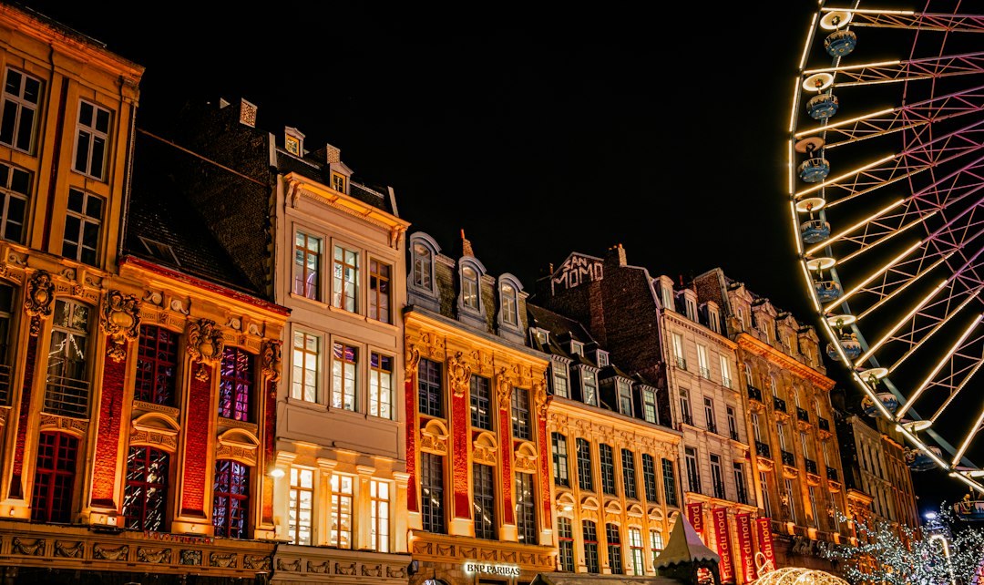 Row of ornate stone building fronts lit orange at night beside a large illuminated ferris wheel