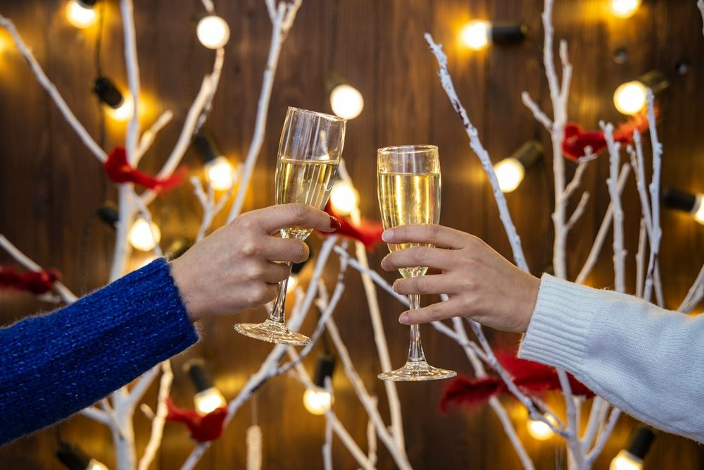 Two hands clinking champagne glasses in front of warm festive lights and white branches